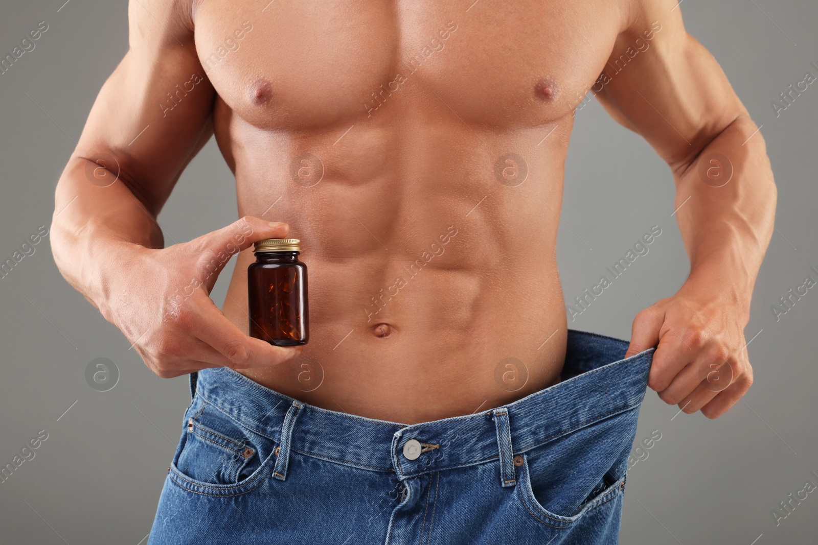 Photo of Athletic man with bottle of supplements wearing big jeans on grey background, closeup. Weight loss