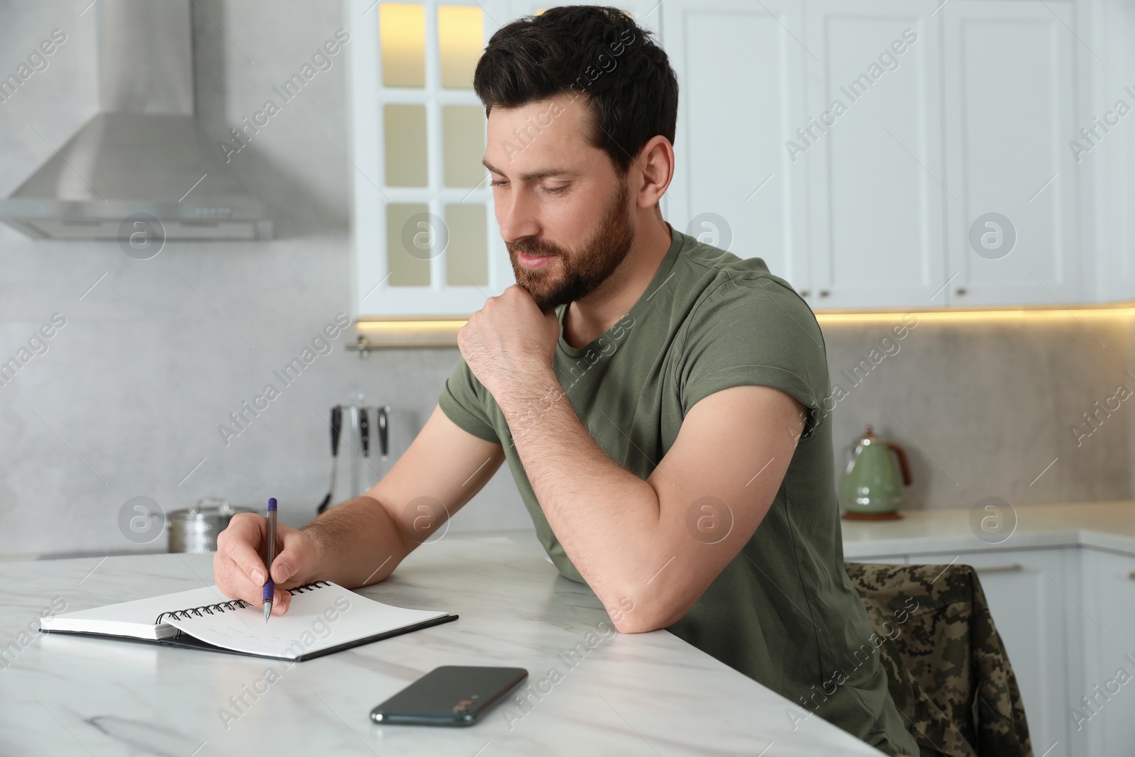 Photo of Soldier writing in notebook at white marble table in kitchen. Military service