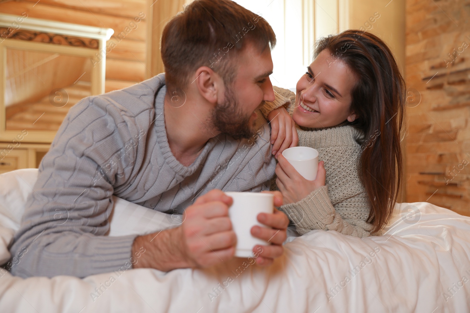 Photo of Young couple in warm sweaters with cups of hot drink on bed at home