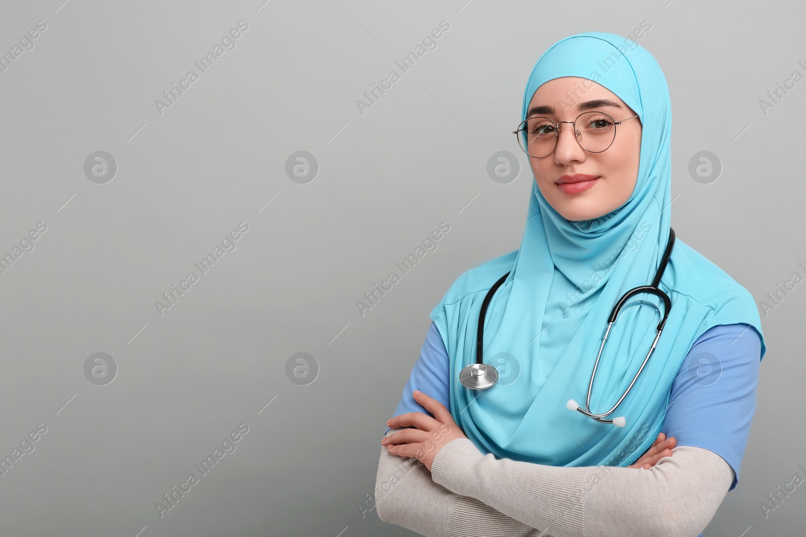 Photo of Muslim woman wearing hijab and medical uniform with stethoscope on light gray background, space for text