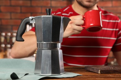 Man with mug of drink at wooden table indoors, focus on coffee moka pot