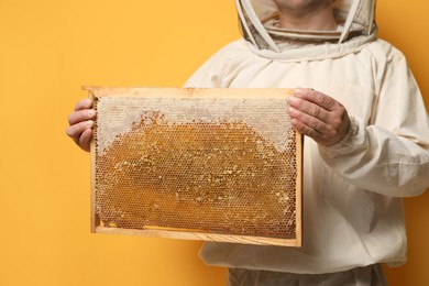 Photo of Beekeeper in uniform holding hive frame with honeycomb on yellow background, closeup