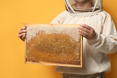 Beekeeper in uniform holding hive frame with honeycomb on yellow background, closeup