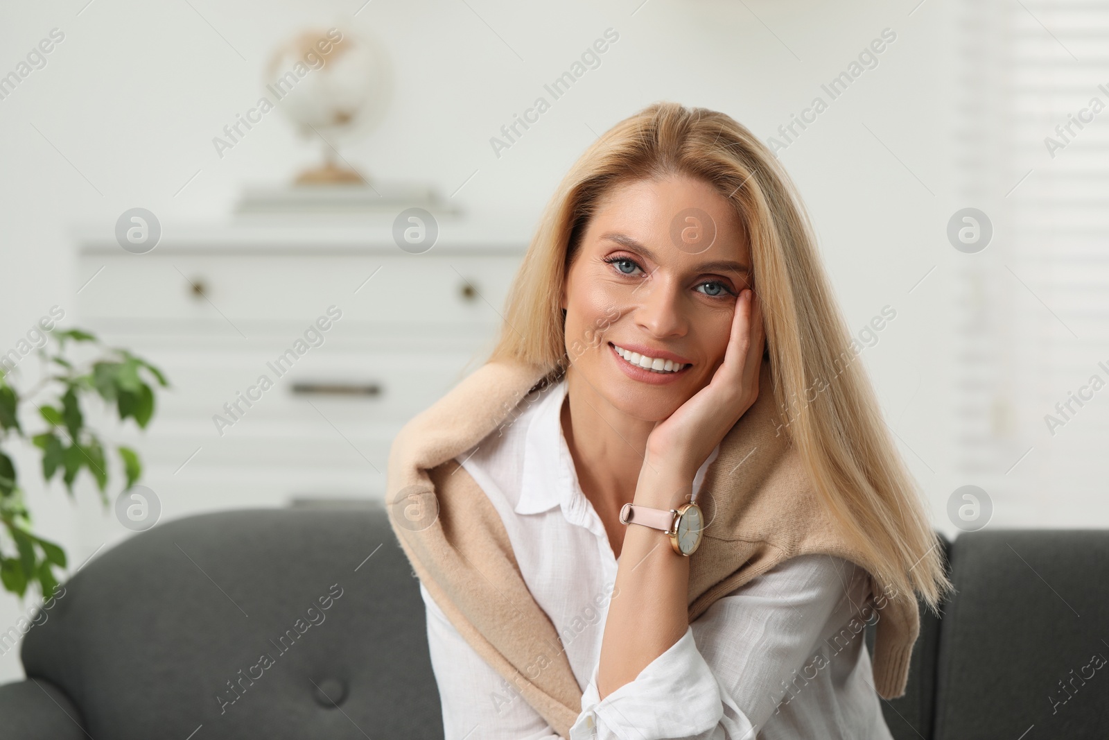 Photo of Portrait of smiling middle aged woman with blonde hair sitting on couch indoors. Space for text