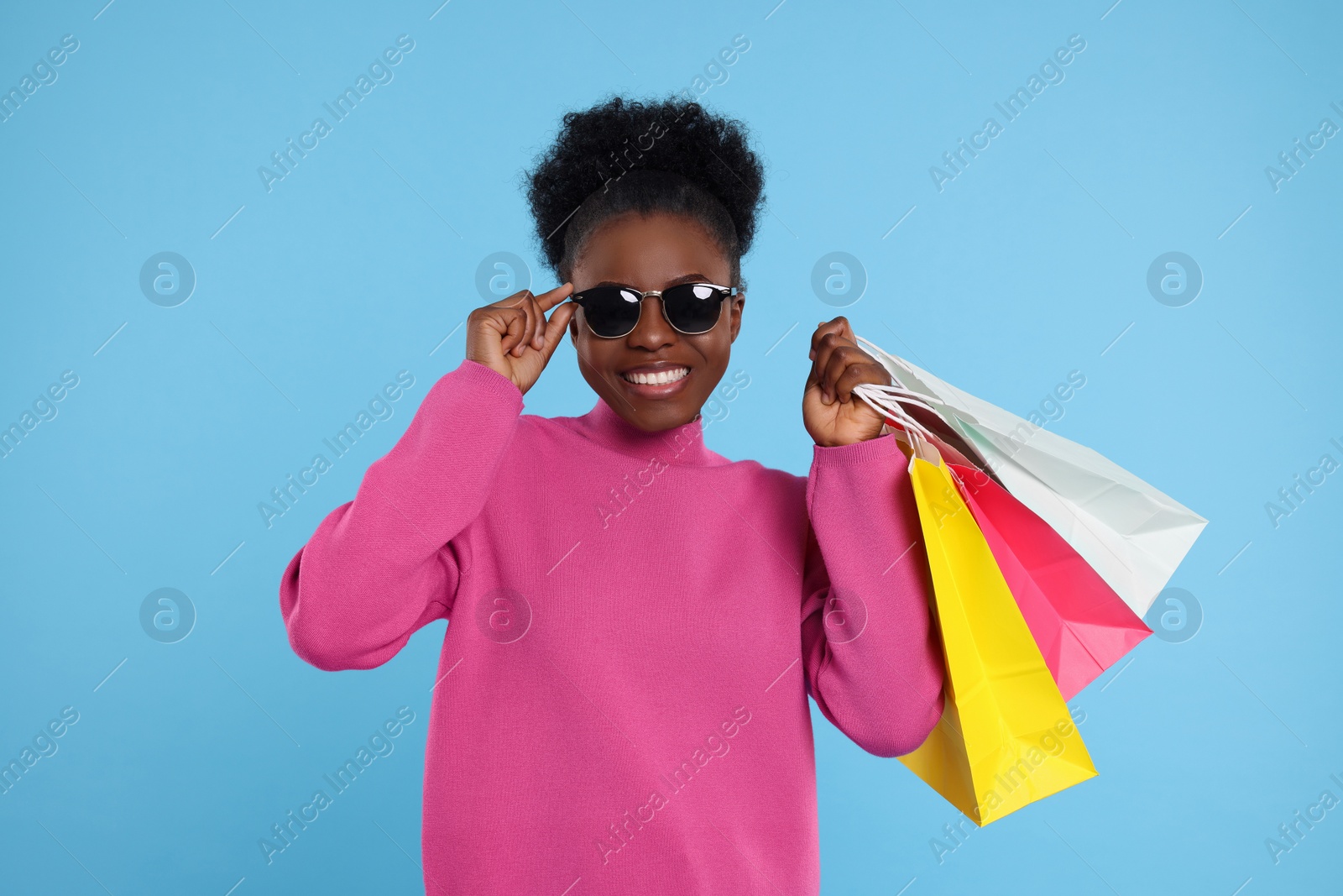 Photo of Happy young woman in stylish sunglasses with shopping bags on light blue background