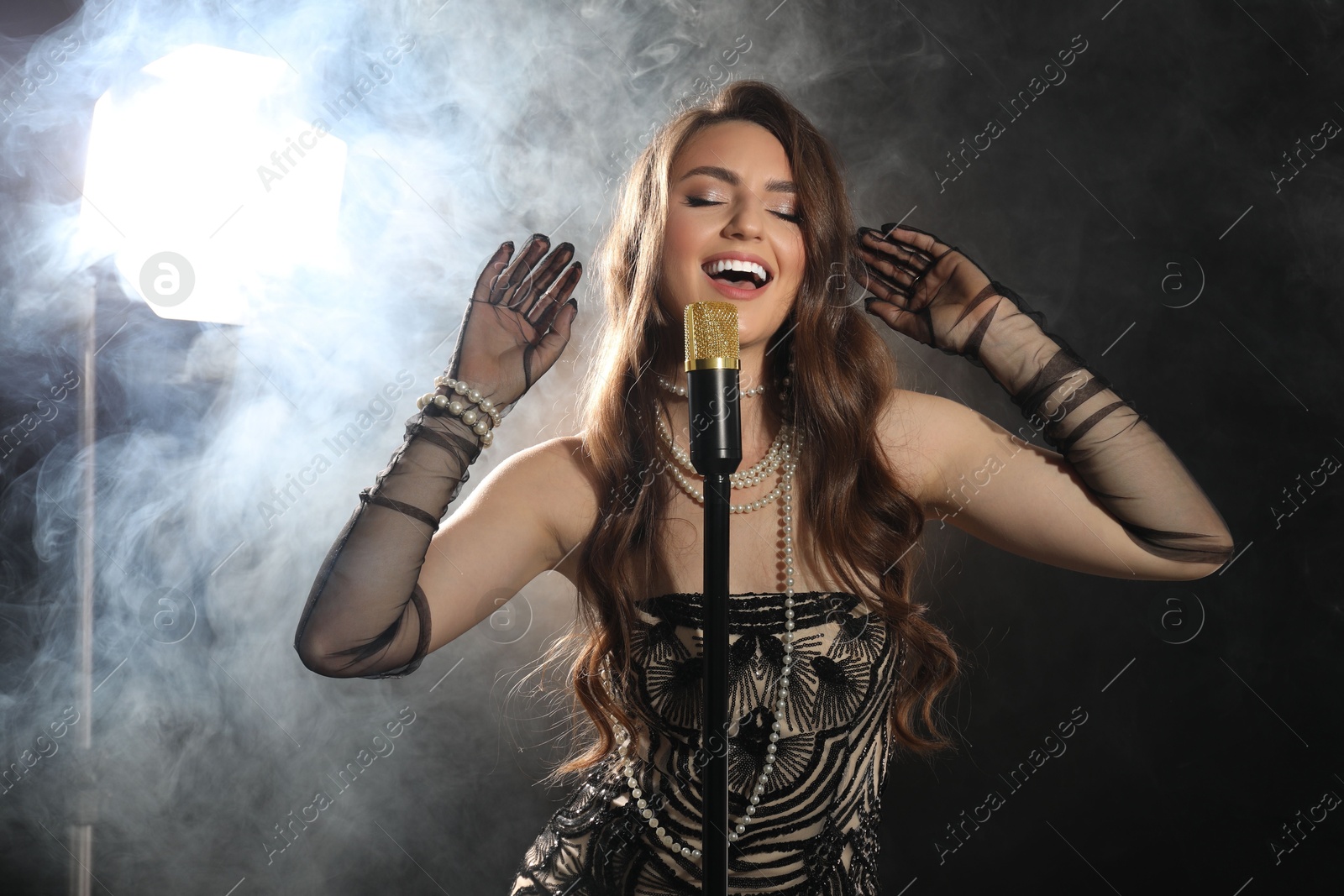 Photo of Beautiful young woman with microphone singing on dark background with smoke