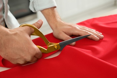 Professional tailor cutting red fabric with scissors in workshop, closeup