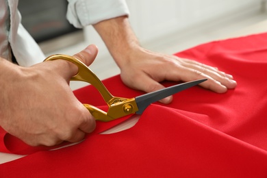 Photo of Professional tailor cutting red fabric with scissors in workshop, closeup