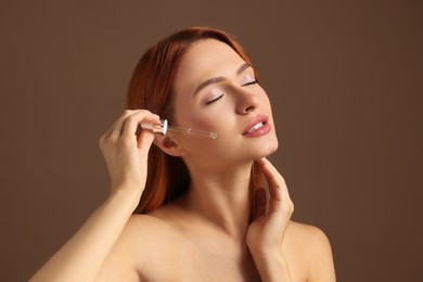 Beautiful young woman applying cosmetic serum onto her face on brown background