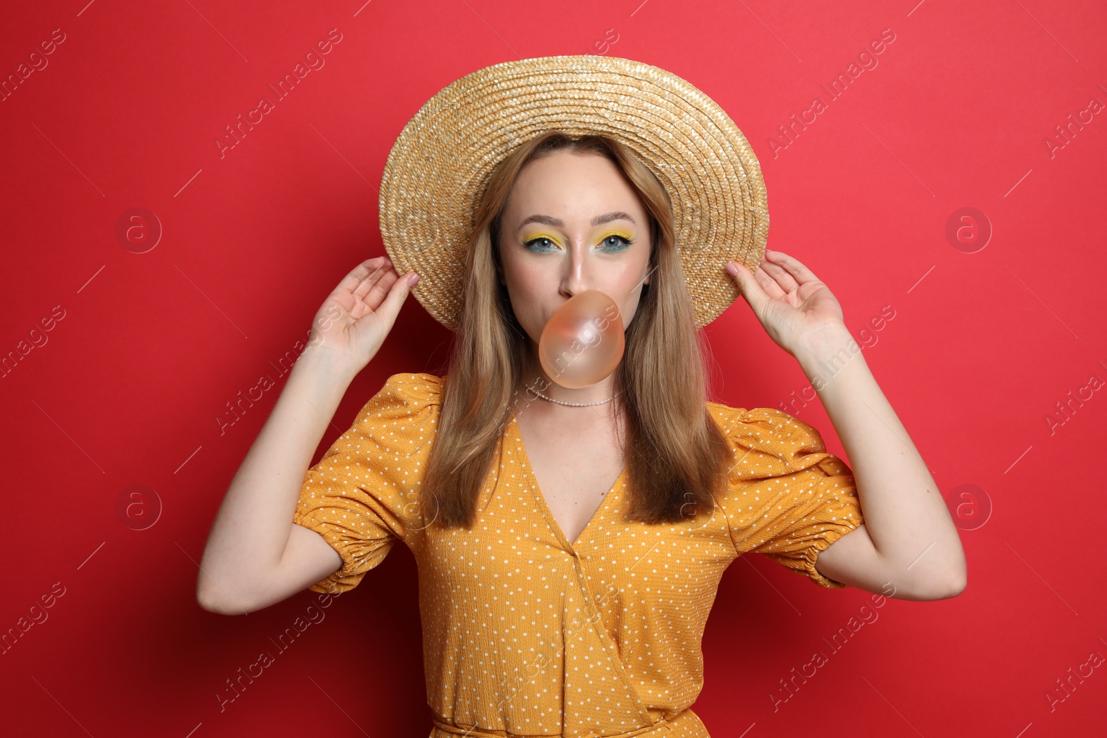 Photo of Fashionable young woman with bright makeup blowing bubblegum on red background