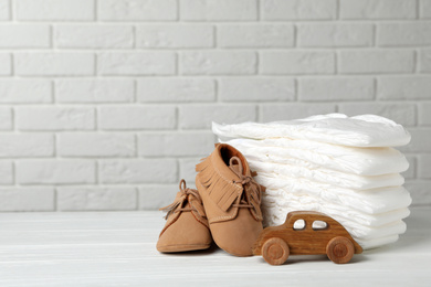 Baby diapers, toy car and child's shoes on wooden table against white brick wall. Space for text