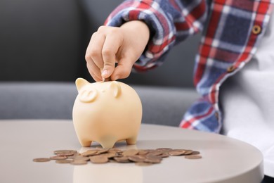 Little boy putting coin into piggy bank at table indoors, closeup