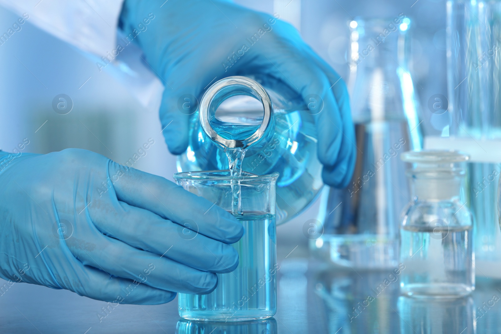 Photo of Doctor pouring blue liquid into beaker from Erlenmeyer flask at grey table, closeup. Laboratory analysis