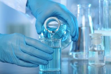 Doctor pouring blue liquid into beaker from Erlenmeyer flask at grey table, closeup. Laboratory analysis