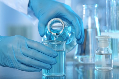 Photo of Doctor pouring blue liquid into beaker from Erlenmeyer flask at grey table, closeup. Laboratory analysis