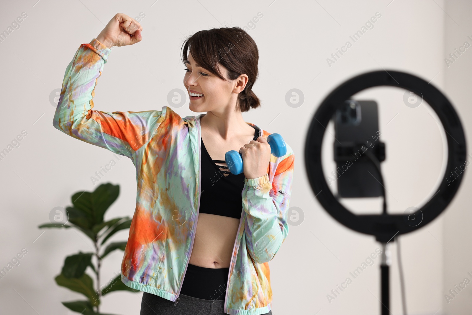 Photo of Happy sports blogger holding dumbbell while streaming online fitness lesson with smartphone at home