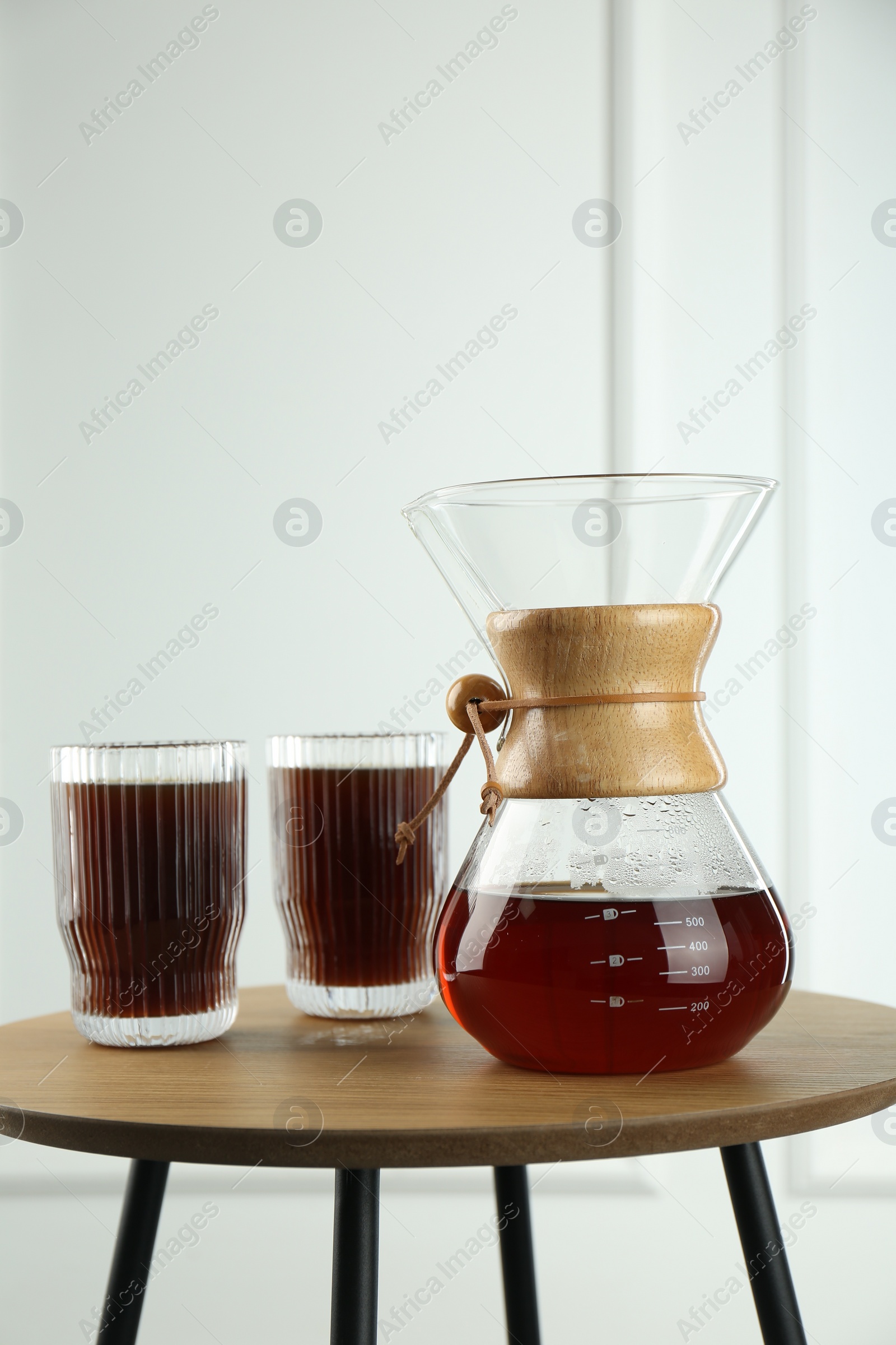 Photo of Glass chemex coffeemaker and glasses of coffee on wooden table against white wall