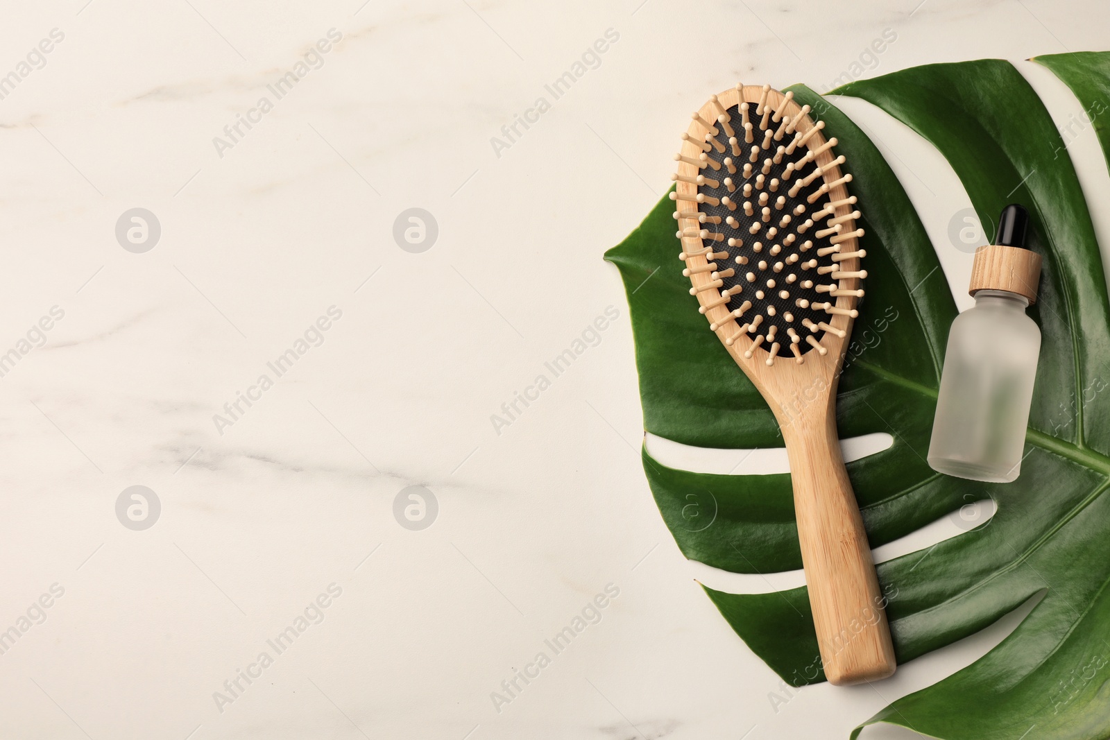 Photo of Wooden hair brush, bottle of essential oil and monstera leaf on white marble table, top view. Space for text