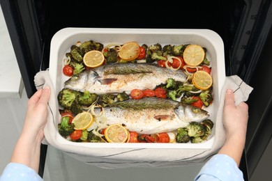 Woman taking baking dish with delicious fish and vegetables from oven, top view