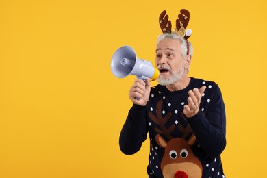 Photo of Senior man in Christmas sweater and reindeer headband shouting in megaphone on orange background. Space for text
