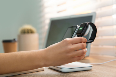 Photo of Woman putting smartwatch onto wireless charger at wooden table, closeup. Modern workplace accessory
