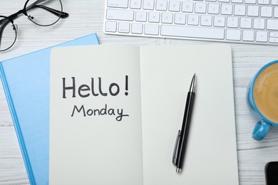 Photo of Message Hello Monday written in notebook, computer keyboard and cup of coffee on white wooden desk, flat lay