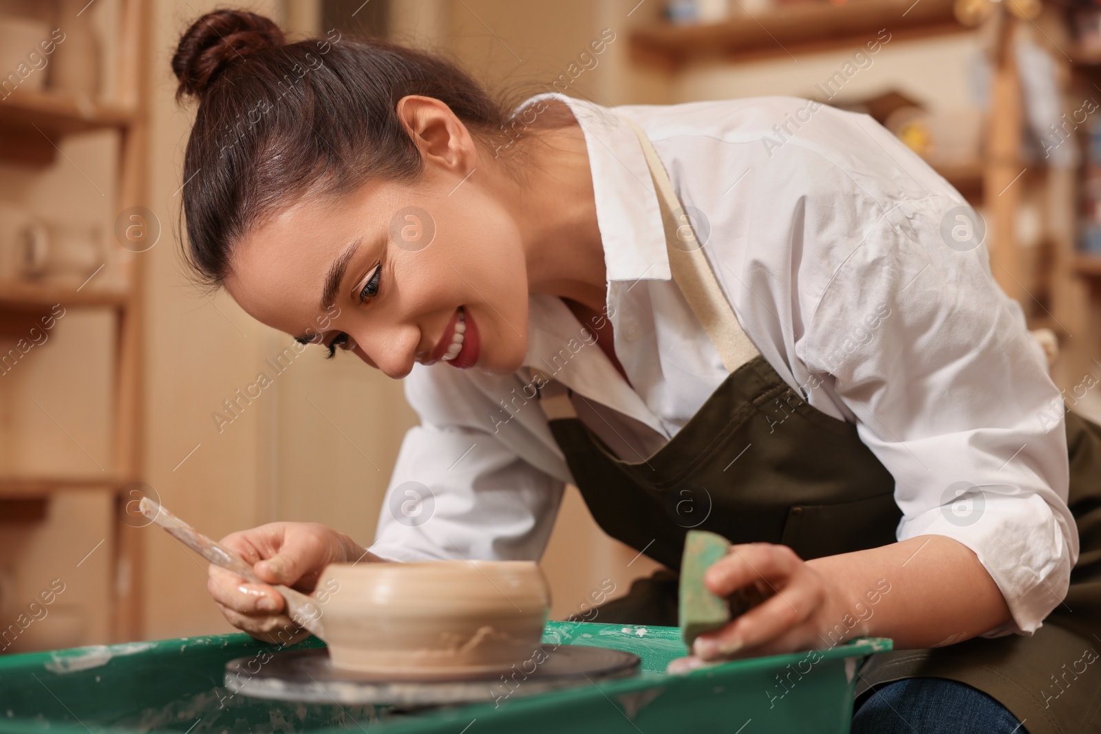 Photo of Clay crafting. Smiling woman making bowl with modeling tool on potter's wheel in workshop