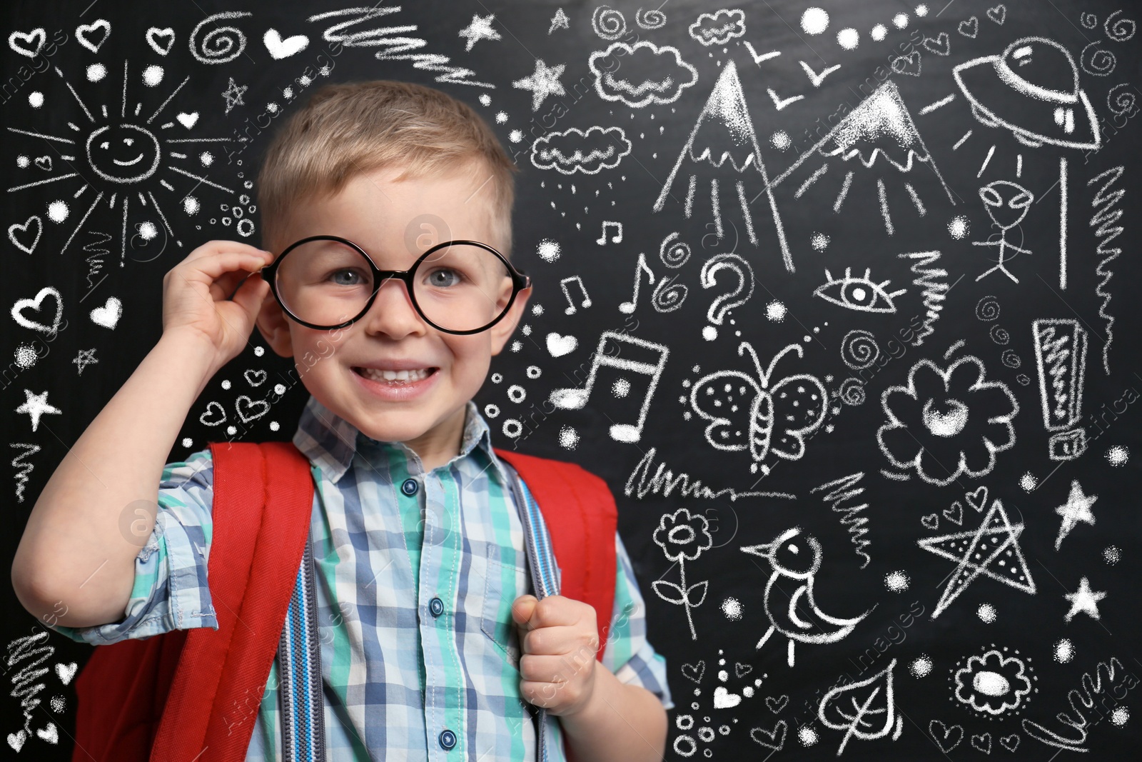 Image of Cute little child in glasses near chalkboard with different drawings. First time at school