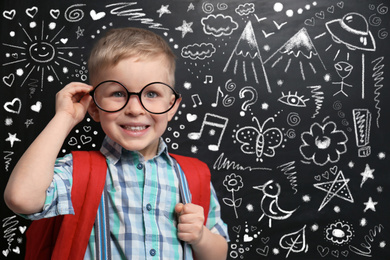 Image of Cute little child in glasses near chalkboard with different drawings. First time at school
