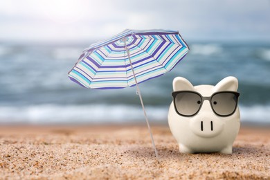 Image of Vacation savings. Piggy bank with sunglasses and umbrella on sandy beach near sea