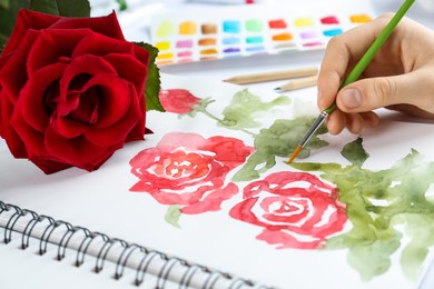 Photo of Woman painting roses in sketchbook at white table, closeup