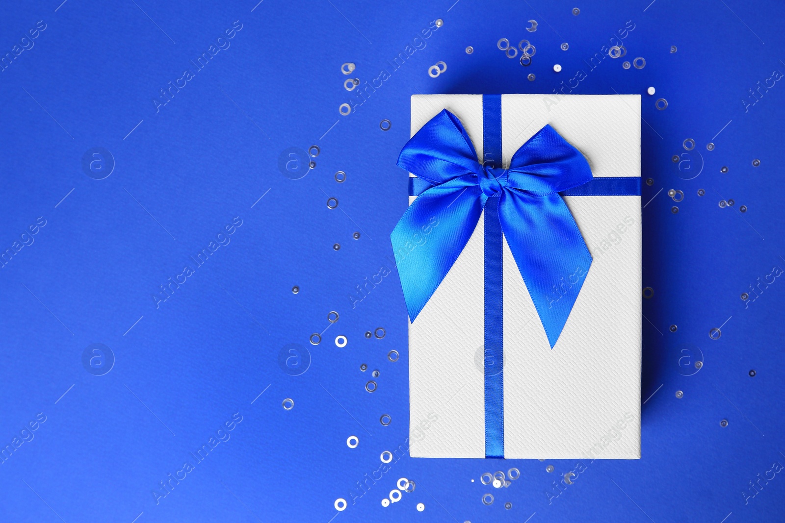 Photo of Beautiful gift box with bow and confetti on blue background, top view. Space for text