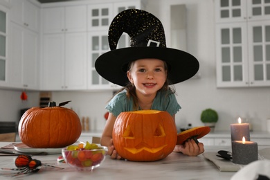 Photo of Little girl with pumpkin jack o'lantern at table in kitchen. Halloween celebration