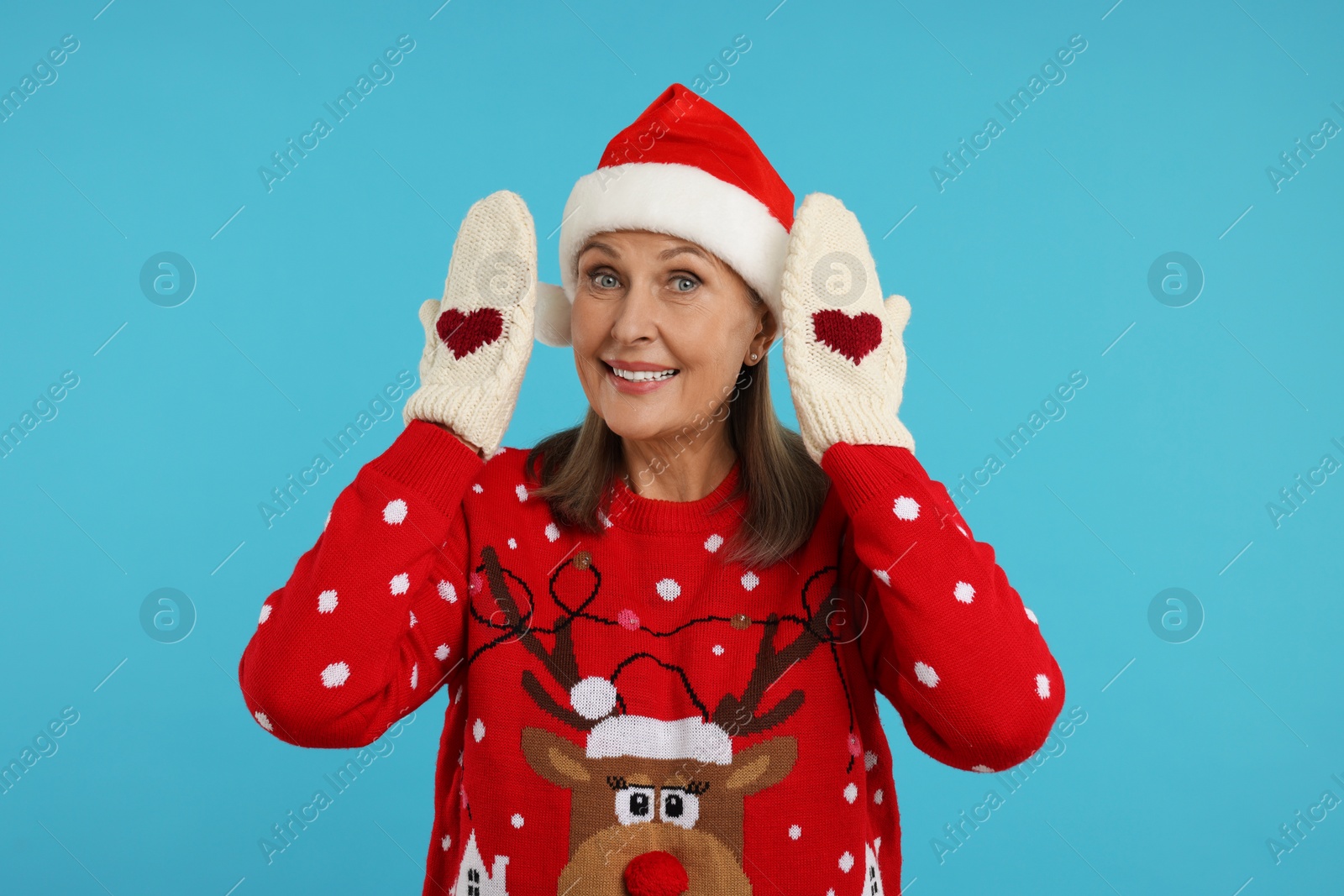 Photo of Senior woman in Christmas sweater, Santa hat and knitted mittens on light blue background