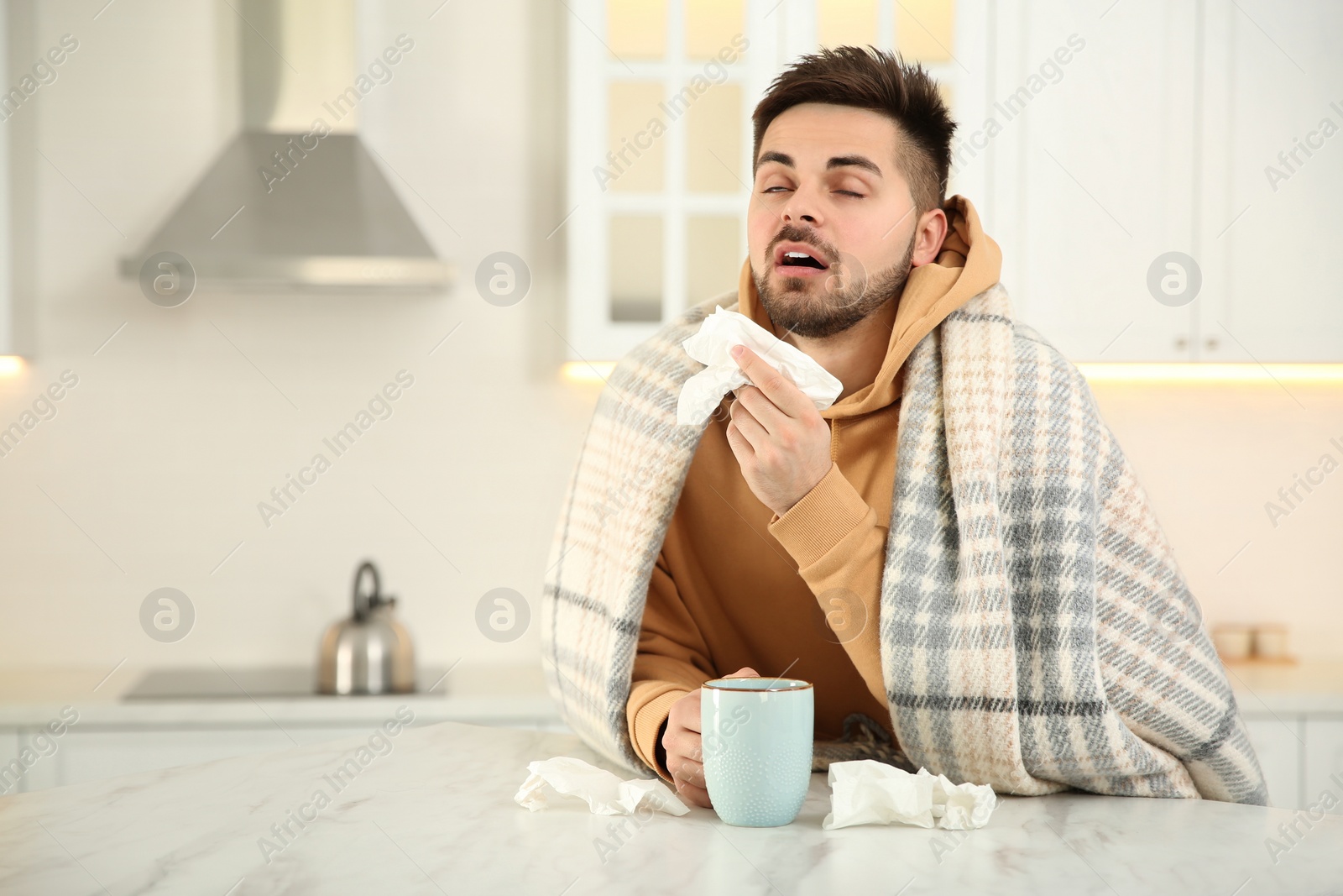 Photo of Sick young man with cup of hot drink and tissues in kitchen. Influenza virus