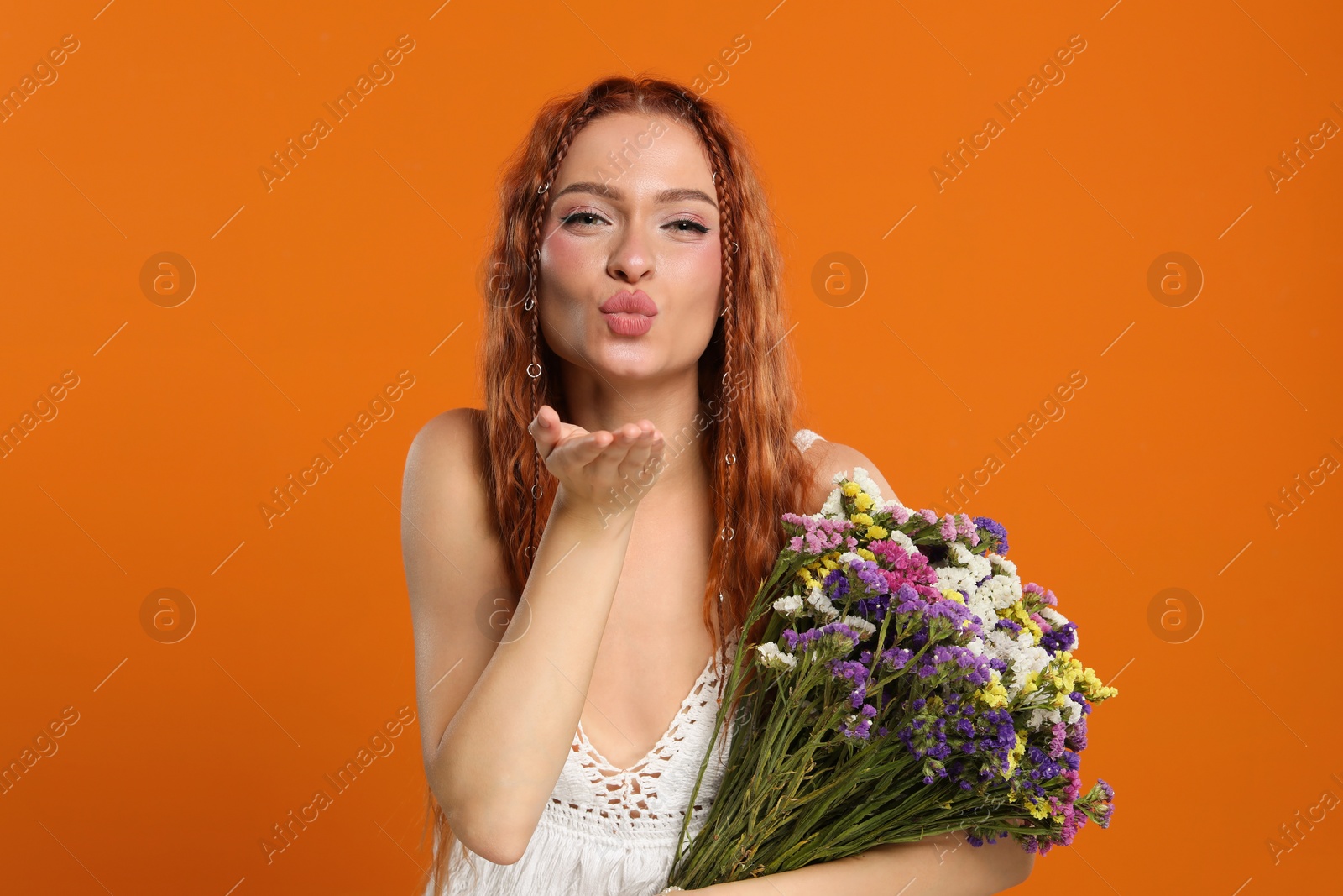 Photo of Beautiful young hippie woman with bouquet of colorful flowers blowing kiss on orange background