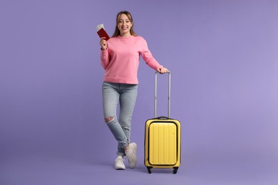 Photo of Happy young woman with passport, ticket and suitcase on purple background