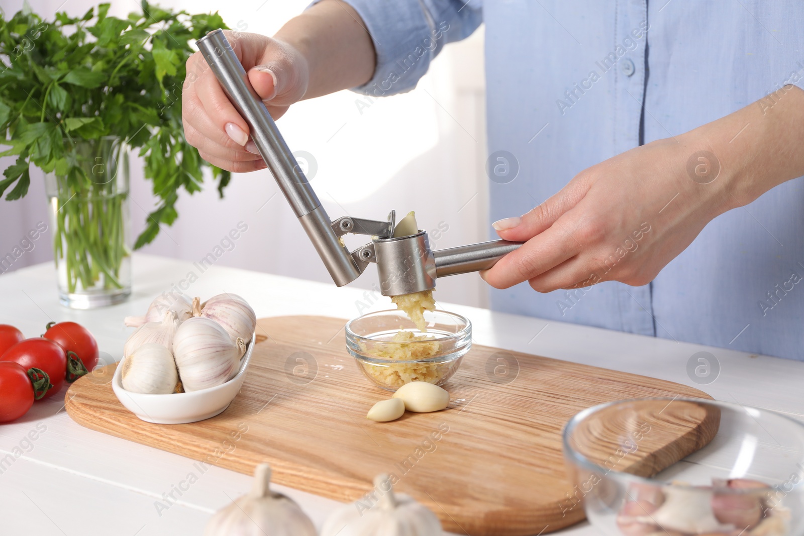 Photo of Woman squeezing garlic with press at white wooden table in kitchen, closeup