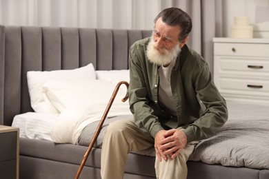 Photo of Senior man with walking cane suffering from knee pain on bed at home. Rheumatism symptom