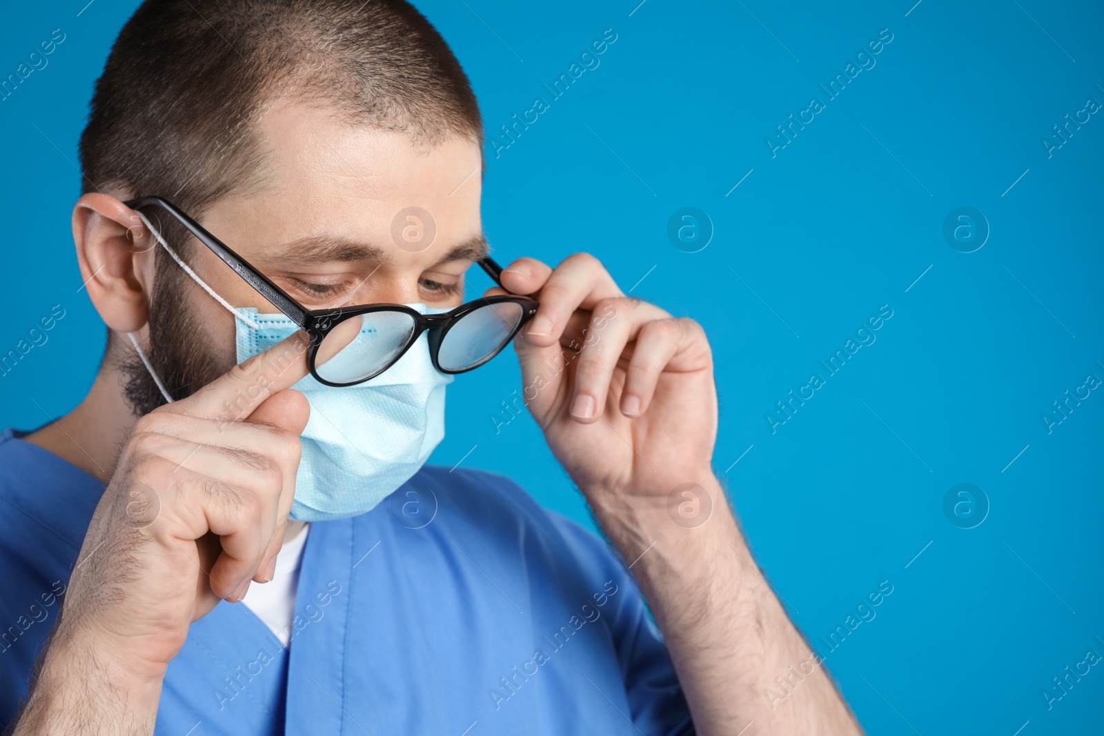 Photo of Doctor wiping foggy glasses caused by wearing disposable mask on blue background. Protective measure during coronavirus pandemic