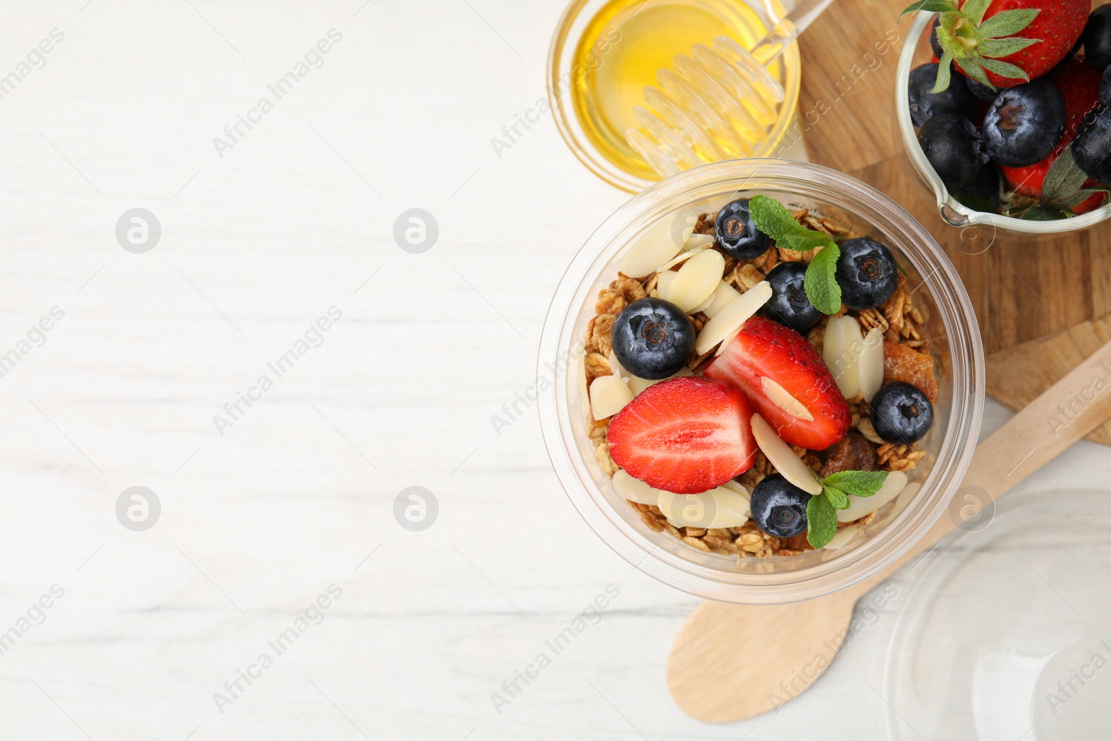 Photo of Tasty granola with berries, almond flakes and mint in plastic cup on white wooden table, flat lay. Space for text