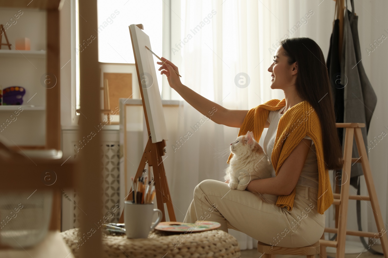 Photo of Beautiful young woman drawing on easel with cat at home