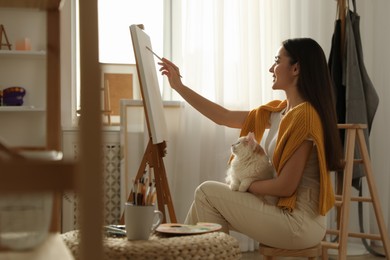 Photo of Beautiful young woman drawing on easel with cat at home