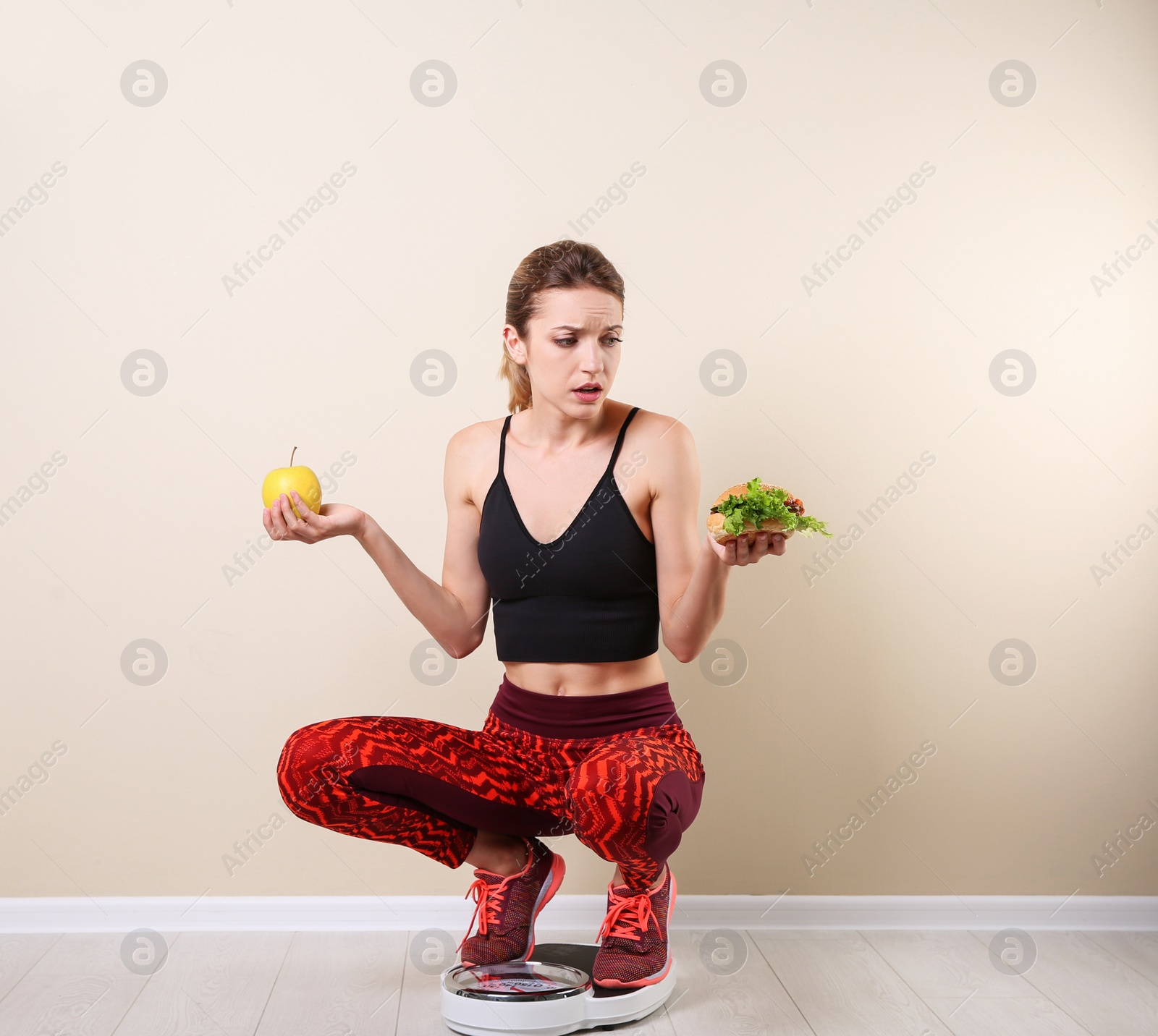 Photo of Woman holding tasty sandwich and apple while measuring her weight on floor scales near light wall. Weight loss motivation