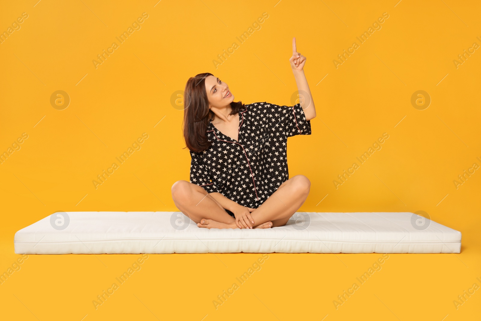 Photo of Young woman sitting on soft mattress and pointing upwards against orange background