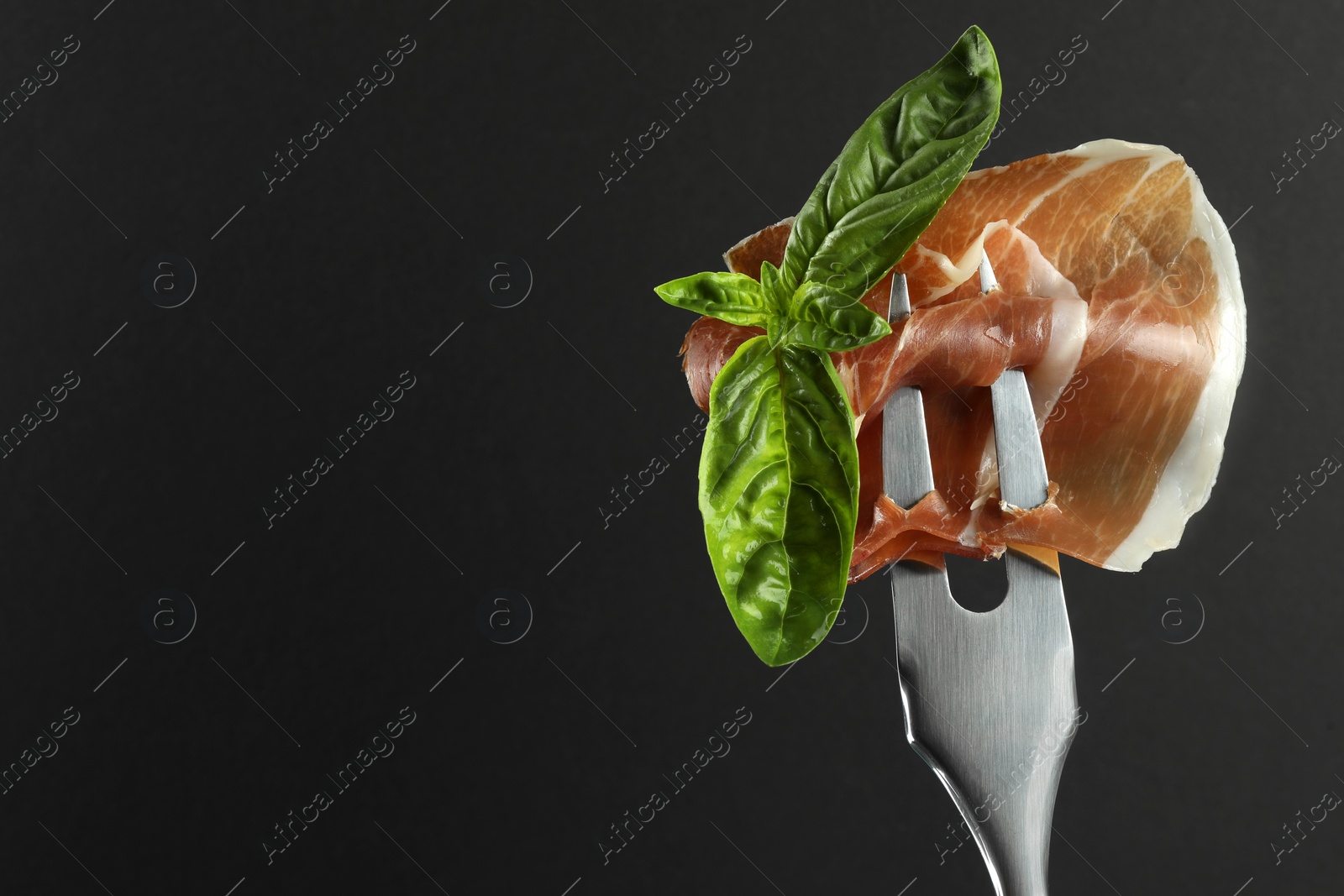 Photo of Fork with slice of tasty jamon and basil leaves on black background, closeup. Space for text