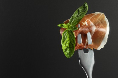 Fork with slice of tasty jamon and basil leaves on black background, closeup. Space for text