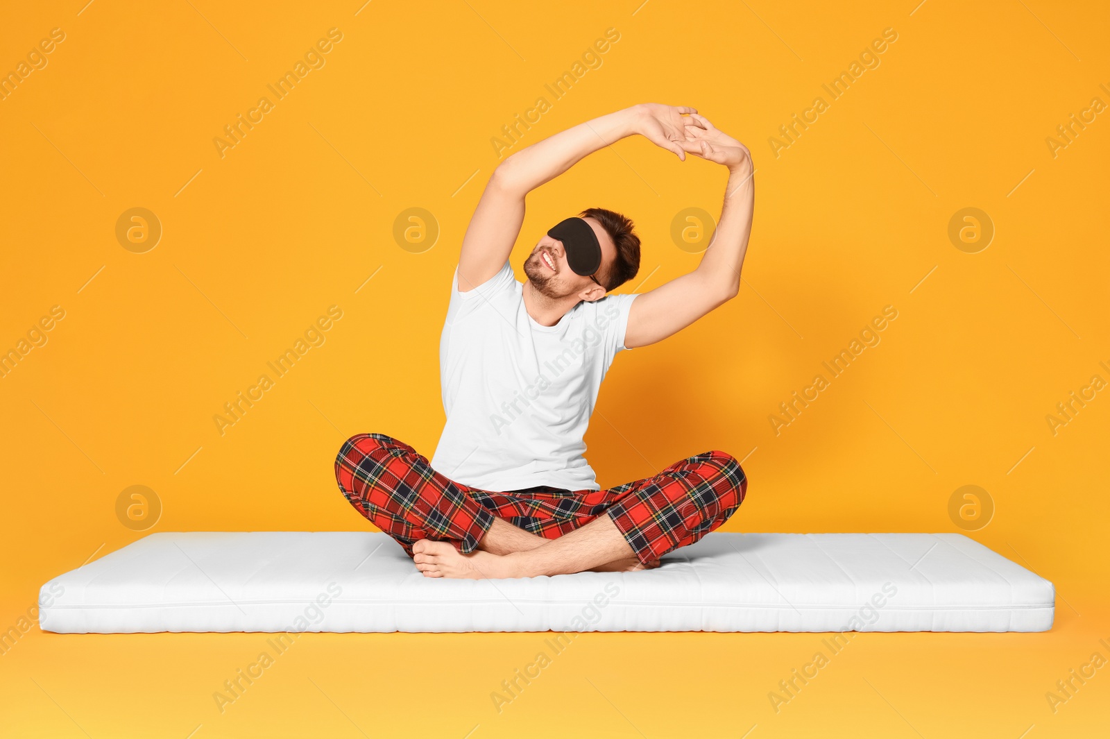 Photo of Man in sleeping mask sitting on soft mattress and stretching against orange background