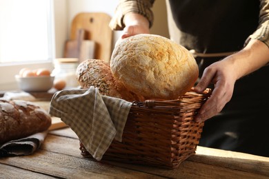 Photo of Man holding wicker basket with different types of bread at wooden table indoors, closeup