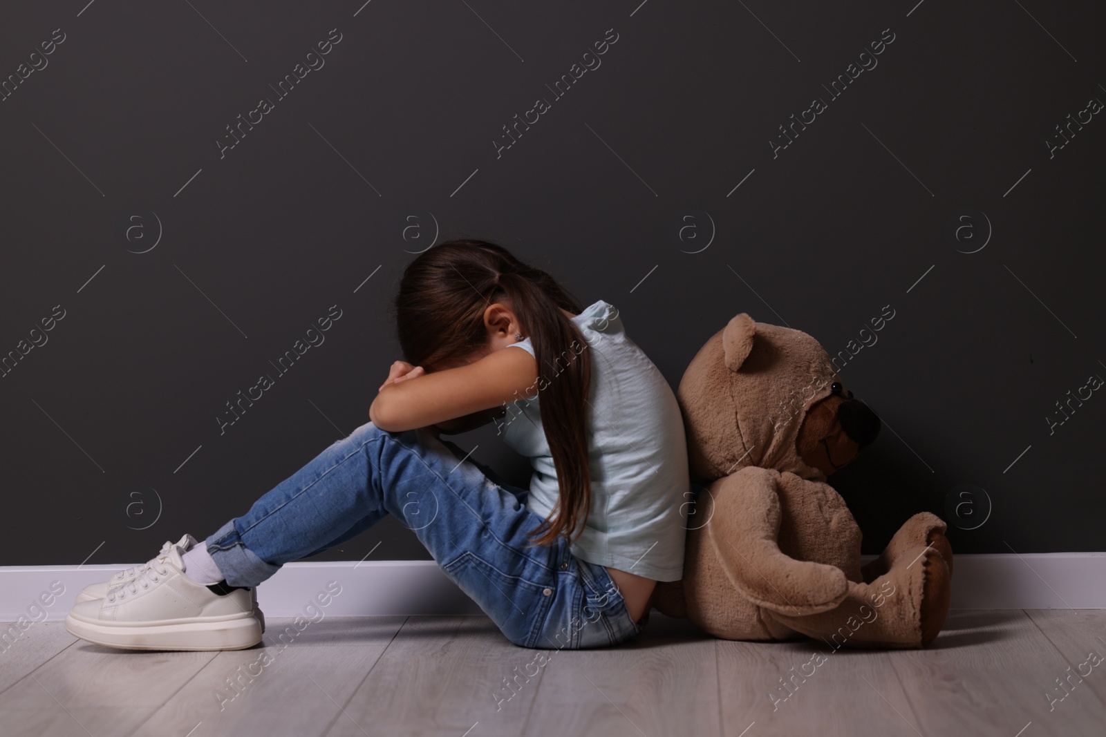 Photo of Child abuse. Upset little girl with teddy bear sitting on floor near gray wall indoors
